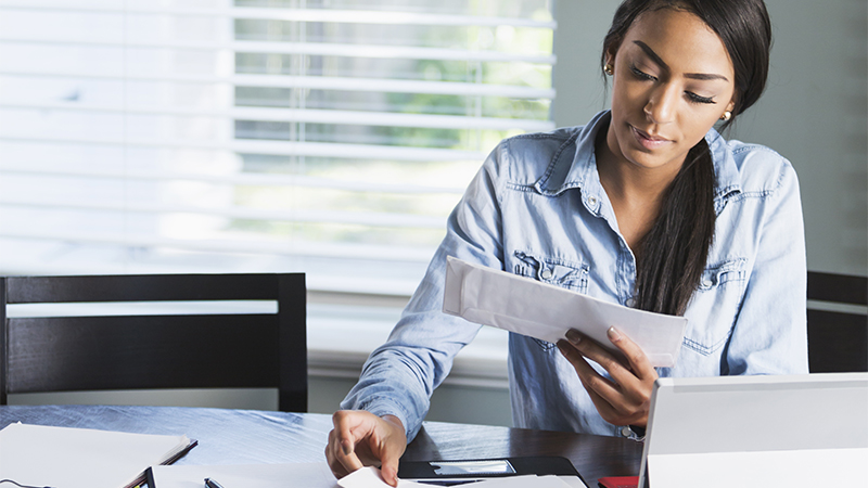 Uma jovem mulher morena está sentada a uma mesa de trabalho, segurando com a mão esquerda uma carta que contempla. A mão direita segura um papel que está pousado na mesa, sobre a qual repousa muita outra papelada e um computador do tipo "phablet". Ao lado da mulher, uma cadeira vazia, e atrás uma grande janela com a persiana meio corrida.